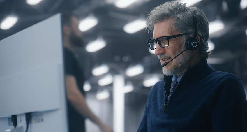 An older man with gray hair and glasses, wearing a headset and dark sweater, sits at a computer workstation in a modern, industrial-style office. Blurred lights and a standing person are visible in the background.