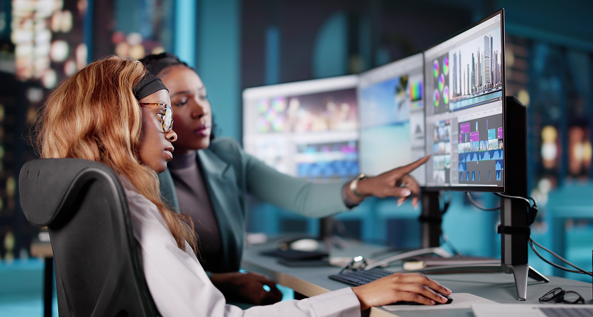 Two women sit at a desk in a modern office, focused on dual monitors displaying video editing software. One woman points at the screen while the other attentively follows, both collaborating on a digital project with city lights blurred in the background.