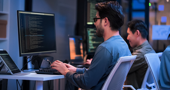 Two people sit at desks with large monitors displaying code in a modern office. The person in the foreground types on a keyboard, wearing glasses and a blue shirt. The workspace appears organized, with additional screens and soft lighting.