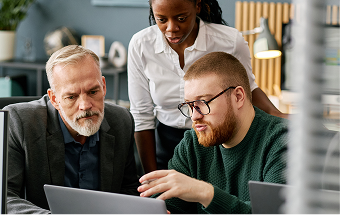 Three colleagues, two men and one woman, gather around a laptop in an office. The bearded man in glasses gestures at the screen, explaining something, while the older man with gray hair and the woman in a white blouse attentively watch and listen.