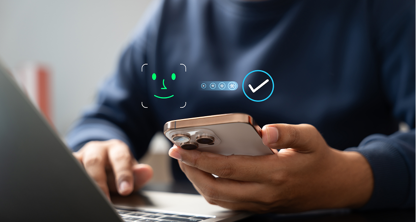A person in a dark blue shirt holds a smartphone in one hand and types on a laptop with the other. Digital facial recognition icons and a checkmark appear, indicating successful facial authentication on the phone. The background is blurred.