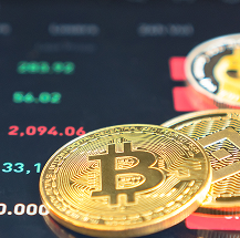 Close-up of a shiny gold Bitcoin coin in focus, with another cryptocurrency coin partly visible behind it. In the background, a digital screen displays fluctuating cryptocurrency prices in green and red numbers, suggesting market activity.