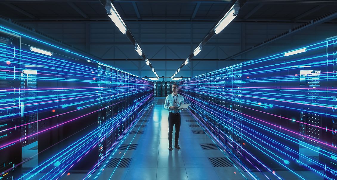 A man stands in a modern server room lined with racks of servers on both sides. Blue and pink light trails, digitally added, represent data transmission, creating a futuristic atmosphere. He holds a tablet and appears to be monitoring or managing the systems.