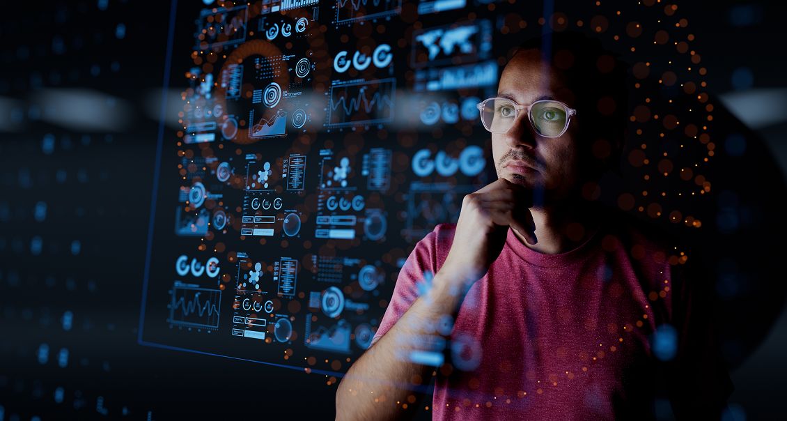 A man in glasses and a red shirt looks thoughtfully at digital data visualizations projected in front of him, featuring charts, graphs, and infographics with blue and orange highlights, suggesting data analysis or technology-focused work.