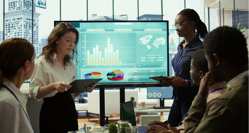 Five professionals in a modern office analyze bar and pie charts on a large screen. Two women stand holding tablets and discuss data, while three colleagues sit and listen. City buildings are visible through large windows in the background.