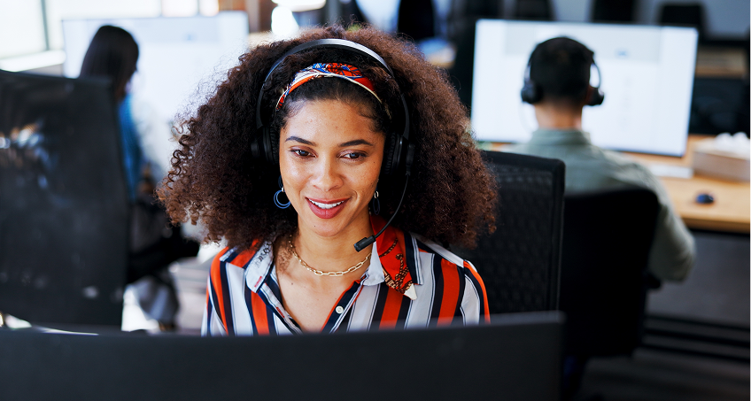 A woman with curly hair, wearing a striped shirt, a headset, and a colorful headband, smiles while working at a computer in an office. Other people wearing headsets work at computers in the background.
