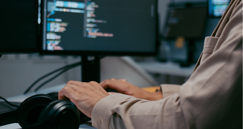 A person wearing a beige jacket types on a keyboard in front of a computer monitor displaying colorful lines of code. A pair of black headphones rests on the desk nearby. The background is blurred, focusing on the hands and screen.