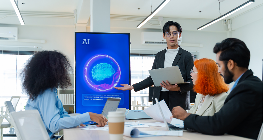 A person stands by a digital screen displaying a blue AI graphic with a brain image, presenting to three seated colleagues. The group discusses documents in a modern office, with laptops, papers, and coffee cups on the table.