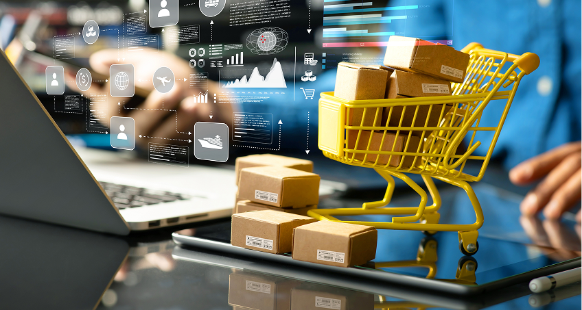 A hand uses a laptop with a stylus near small cardboard boxes in a yellow shopping cart, symbolizing online shopping. Digital icons and graphs hover in the air, reflecting e-commerce, logistics, and data analysis concepts.