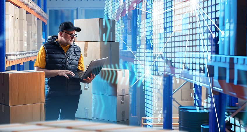 A man in a cap and vest stands in a warehouse among stacked boxes, using a laptop. Blue digital graphics and data visualization overlays highlight technology and inventory management in a modern logistics environment.