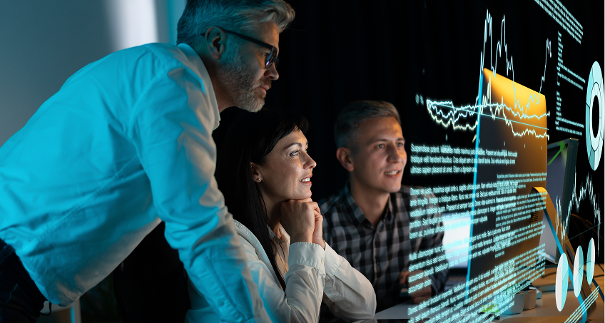 Three people, two men and a woman, are looking intently at a computer screen displaying digital data, charts, and graphs in a dark room with blue lighting, suggesting a late-night work session or data analysis in a tech-focused environment.