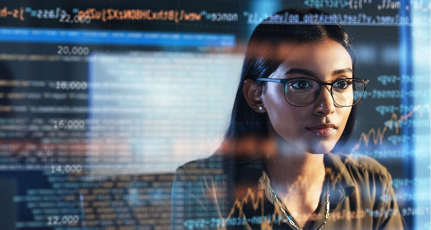 A woman wearing glasses looks intently at a computer screen filled with colorful code and graphs. The coding and data visualizations are visible as reflections, suggesting she is working on programming or data analysis. The setting is tech-focused.