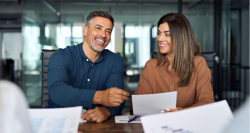 Two people sit at a conference table in a modern office, smiling and holding papers. The man, with short gray hair and a beard, gestures while talking. The woman, with long brown hair, listens and smiles. Paperwork and blurred figures are in the foreground.