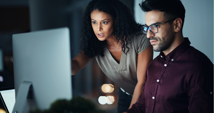 A woman leans over and points at a desktop computer screen while a man in glasses sits and looks at the monitor. Both are focused, working together in a modern office with dim lighting and blurred lights in the background.