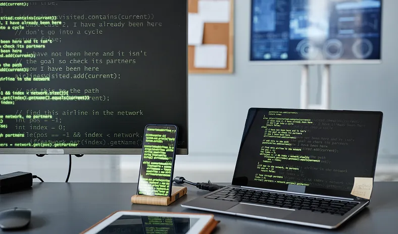 A desktop monitor, laptop, and smartphone display green code on black screens. The devices sit on a modern desk with a blurred office background, including a whiteboard and another screen. The workspace appears organized and tech-focused.