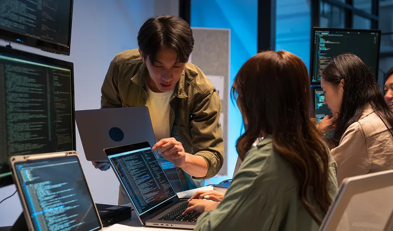 Three people work on laptops with code on multiple monitors in a modern office. One man stands, explaining something to a woman seated at a desk. Another woman in the background types on her laptop. The setting is collaborative and tech-focused.