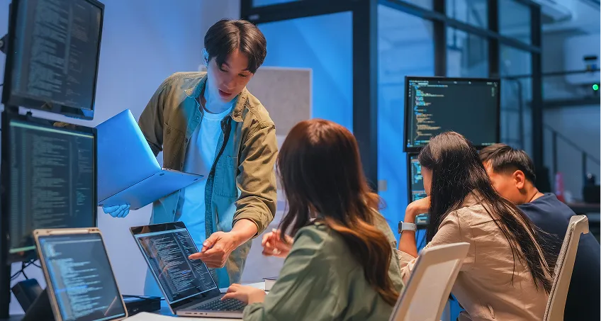 Four people work together in a modern office filled with computer monitors displaying code. One person stands, holding a laptop and pointing at a colleague’s screen, while three others sit at desks, focused on their monitors and collaborating.
