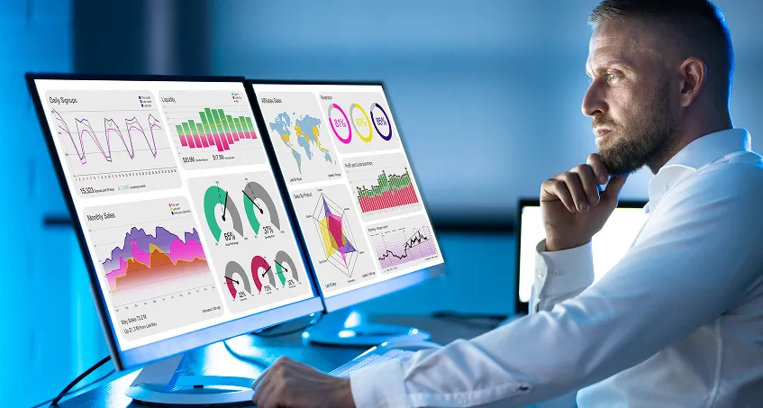 A man in a white shirt sits at a desk, intently viewing two large monitors displaying colorful data visualizations, charts, maps, and graphs in a modern, dimly lit office environment.