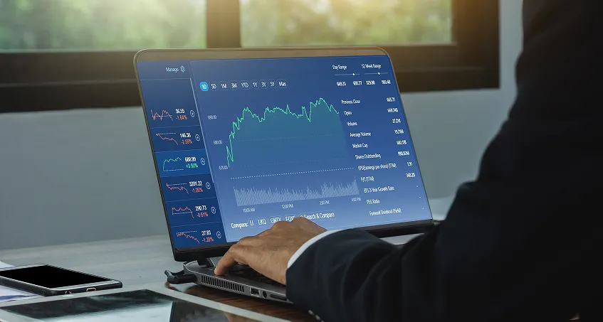 A person in a suit is using a laptop displaying financial charts and stock market data. Multiple graphs with fluctuating lines are shown on the screen. The scene is set in a bright room with a large window and a smartphone lies on the desk nearby.