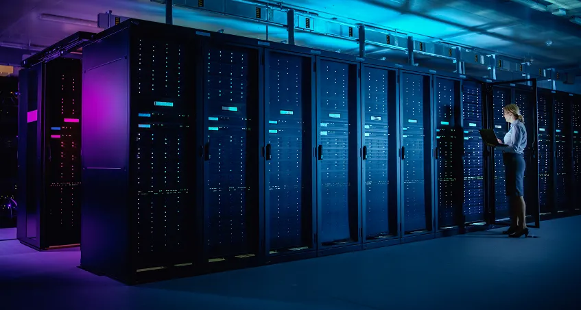A woman stands in a modern server room, working on a laptop. Tall black server racks with glowing blue and purple lights line the room, creating a high-tech atmosphere. The environment is clean and organized, with overhead cables visible.