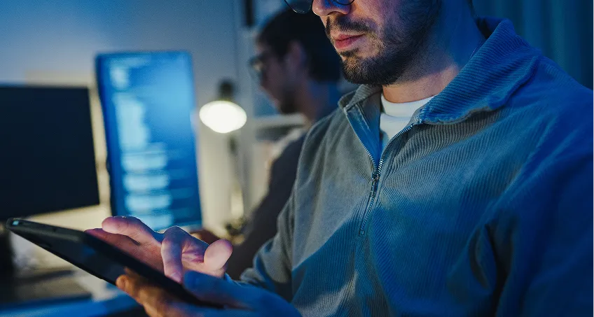A man wearing glasses uses a tablet in a dimly lit room, with another person in the background at a desk. Computer monitors in the background display lines of code, suggesting a tech or programming environment.