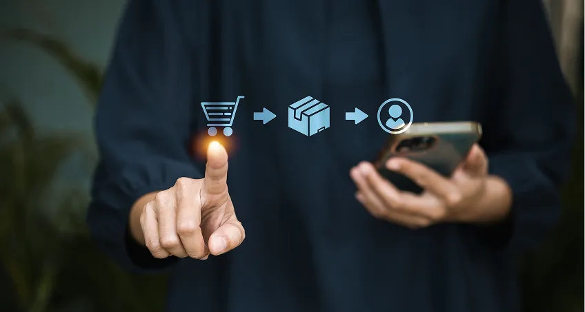 A person taps a virtual shopping cart icon with one finger while holding a smartphone. Icons of a box and a user, connected by arrows, appear in front, representing an online shopping and delivery process. The background is blurred.