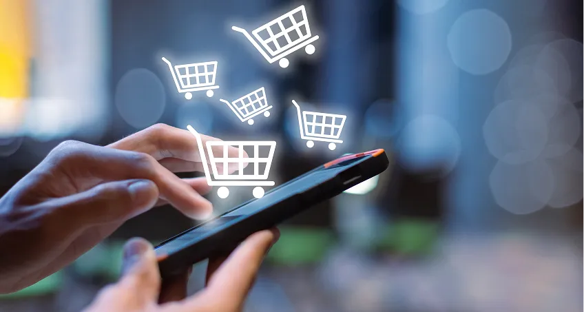 A person uses a smartphone with one hand, while the other hand touches the screen. Several white shopping cart icons appear to float above the phone, symbolizing online shopping. The background is blurred with blue and gray bokeh lights.
