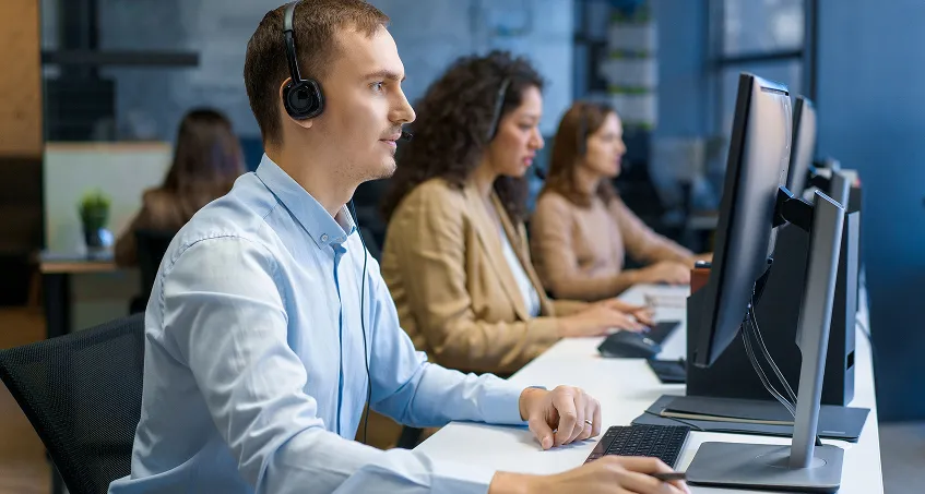 Three people sit at desks in a modern office, each wearing headsets and working on desktop computers. The focus is on a man in a light blue shirt in the foreground, with two women working behind him. The office is bright and organized.