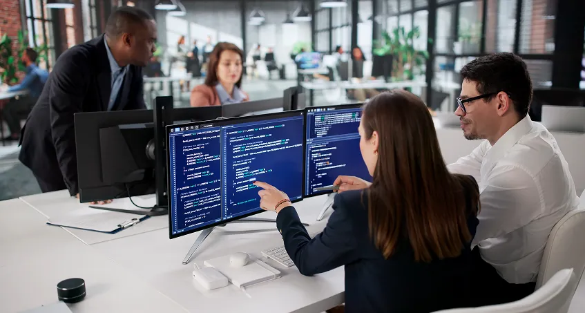 Two colleagues sit at a desk, collaborating and pointing at code on dual monitors. In the background, two more people discuss something near other computer screens in a modern, glass-walled office with green plants and bright lighting.