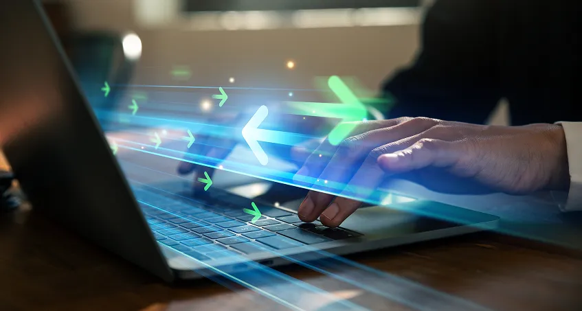 A person’s hands typing on a laptop keyboard. Bright, digital green and blue arrows point right, symbolizing fast data transfer or digital progress. The scene suggests technology, efficiency, or speed in digital work, with a blurred office background.