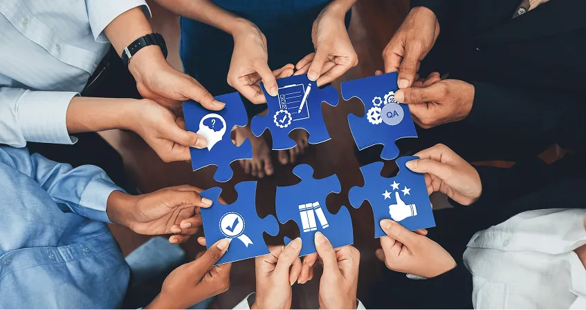 A group of people stands in a circle, each holding a large blue puzzle piece. The pieces display white icons, including a light bulb, briefcase, gears, handshake, checklist, and rating stars, symbolizing teamwork and collaboration.