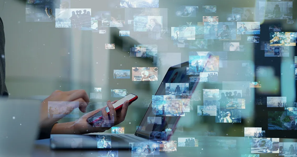 A person uses a smartphone and a laptop at a desk, surrounded by floating digital images and icons, representing multimedia, data transfer, and digital connectivity in a blurred, technology-focused environment.
