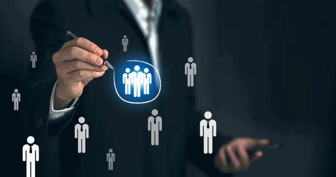 A person in a dark suit holds a pen, drawing a blue circle around a group of three icons resembling people, among other similar icons floating in the air, symbolizing selection or targeting a specific group. The background is blurred and dark.