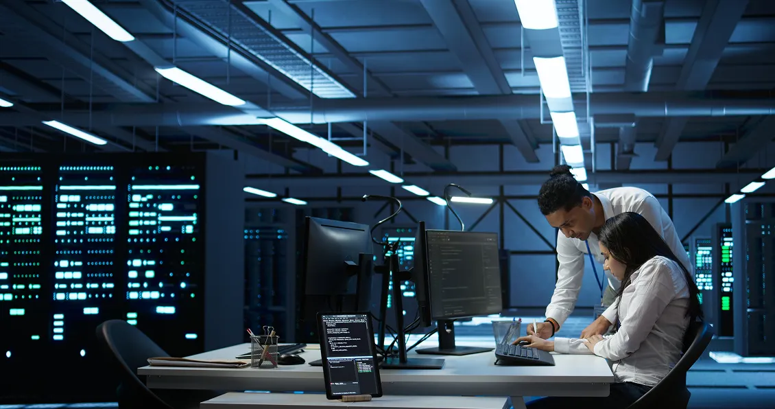 Two professionals work in a modern server room, surrounded by monitors displaying code and network data. One stands, pointing at a document on the desk, while the other sits, focused. Server racks with glowing lights are visible in the background.