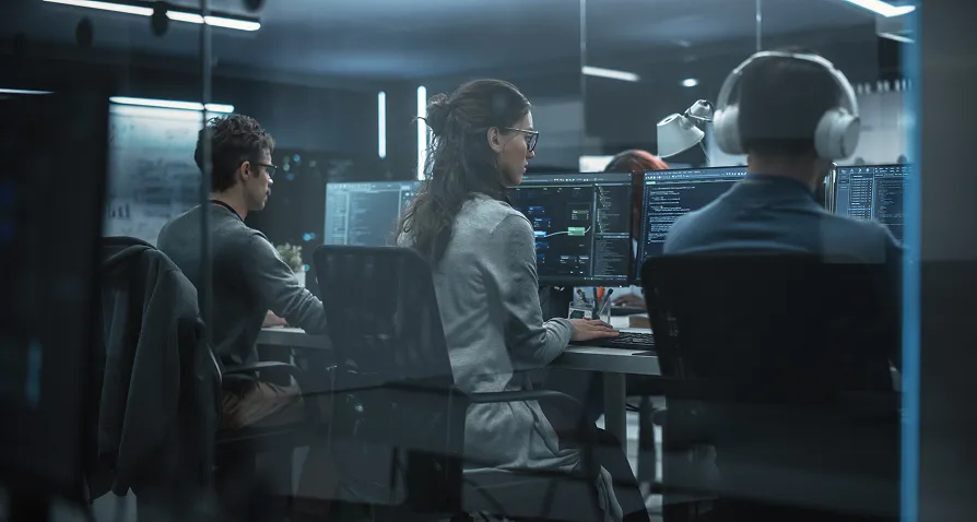 Three people sit at desks in a dimly lit office, focused on computer monitors displaying lines of code. One person wears headphones, another wears glasses, and the atmosphere is serious and professional, suggesting a software development or IT environment.