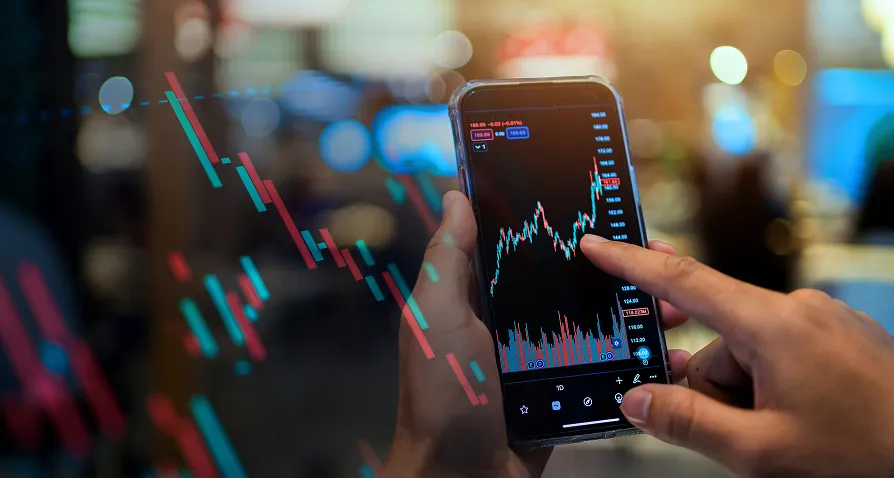 A person holds a smartphone displaying a candlestick stock chart with red and green lines. Their finger touches the screen, analyzing the data. Transparent charts overlay the image, and the blurred background suggests an indoor setting with warm lights.