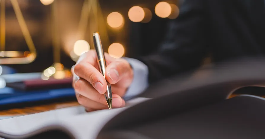 A person in a suit writes with a gold pen on a document at a desk. The image is focused on the hand holding the pen, with blurred legal scales and warm lights in the background, suggesting a professional, legal, or business setting.