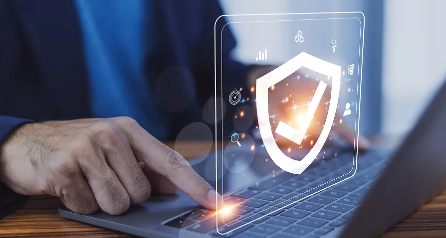 A person’s hand points at a laptop keyboard, with a glowing digital shield and checkmark icon overlay symbolizing cybersecurity. The scene suggests secure technology or data protection, with abstract graphs and icons in the background.