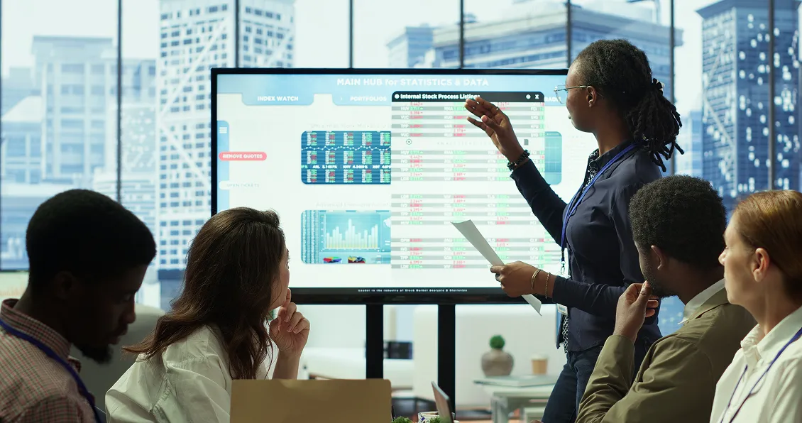 A woman stands and points at a large screen displaying charts and data, while holding papers. Four colleagues, seated around a table, watch and listen. The background shows tall city buildings through large windows.