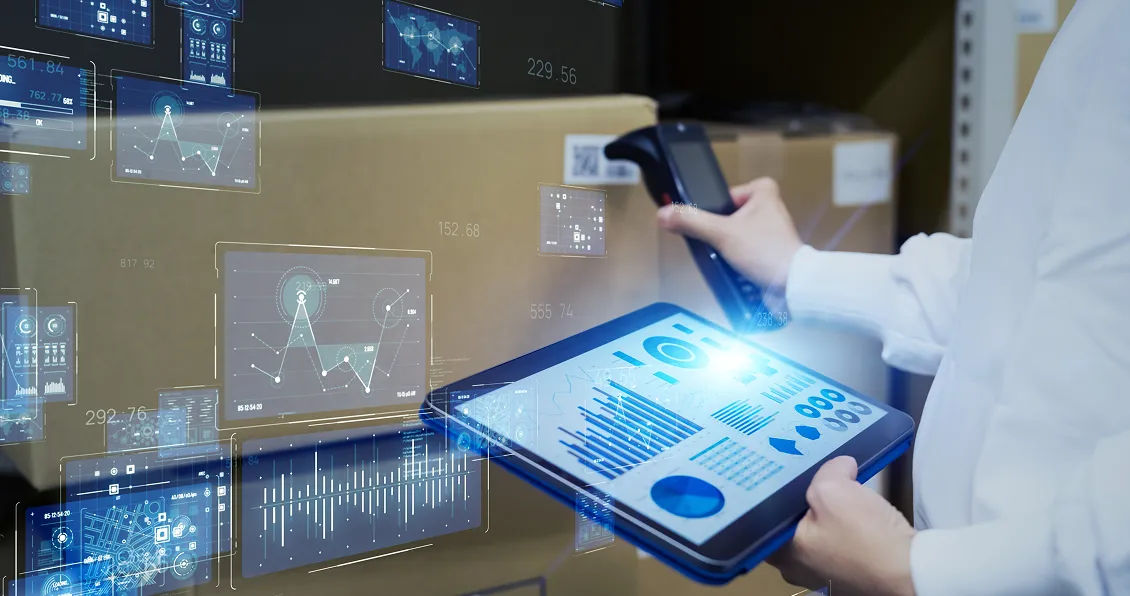 A person in a white coat holds a barcode scanner and a tablet displaying blue digital graphs and charts, with holographic data interfaces superimposed. Cardboard boxes are stacked in the background, suggesting a warehouse or logistics setting.