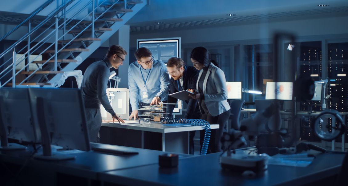 Four people in business attire stand around a table in a modern office or lab, examining electronic components and documents. Computer monitors, cables, and technical equipment are visible; the setting is high-tech and collaborative.