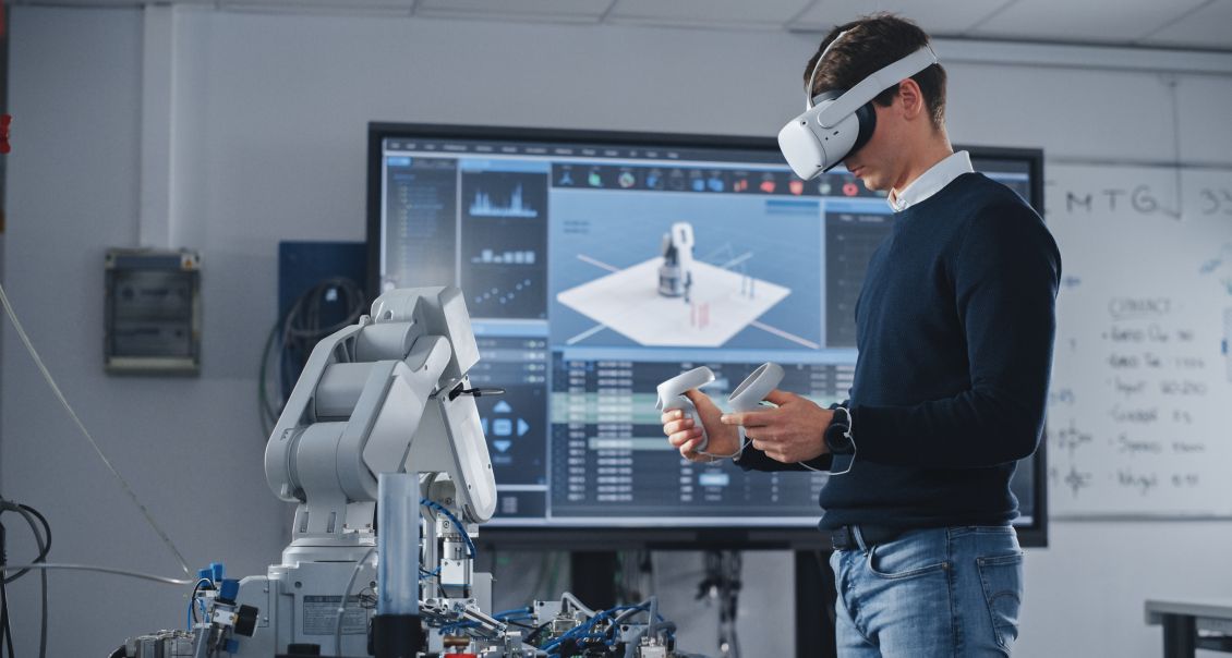 A man wearing a VR headset operates a robotic arm using hand controllers in a high-tech lab. A large monitor behind him displays a 3D model and technical data, and whiteboards with writing are visible on the wall.