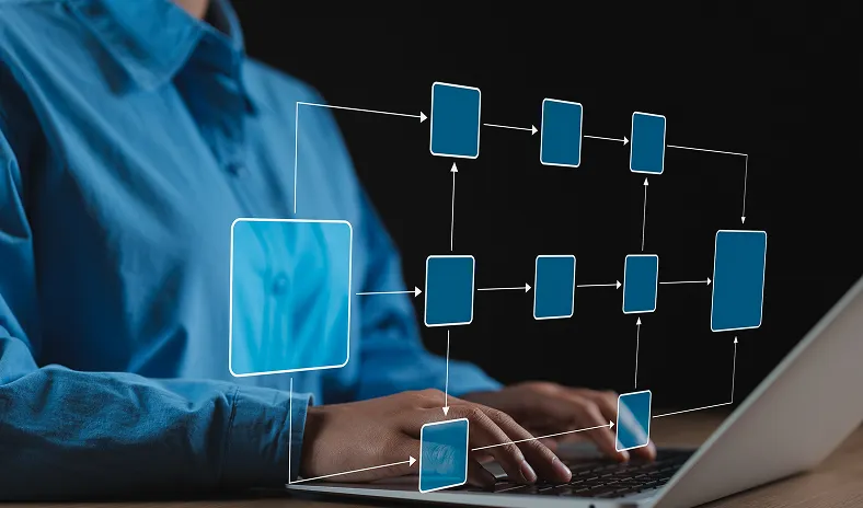 A person in a blue shirt types on a laptop. Overlaid, there is a transparent digital flowchart with interconnected blue rectangles and arrows, symbolizing data flow, processes, or organizational structure, set against a dark background.