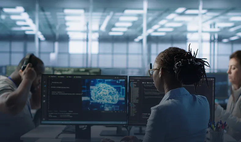 Three people sit at desks with computer monitors displaying code and a digital brain graphic in a modern, glass-walled office. The person in the center, with dark skin and dreadlocks, is focused on their screen. The environment is bright and high-tech.