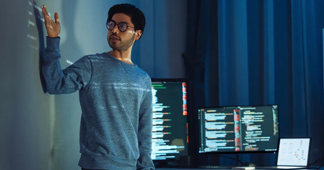 A man wearing glasses and a grey sweater gestures toward a projection on a wall. Behind him are two monitors displaying code and technical diagrams in a dimly lit, modern workspace with blue curtains.