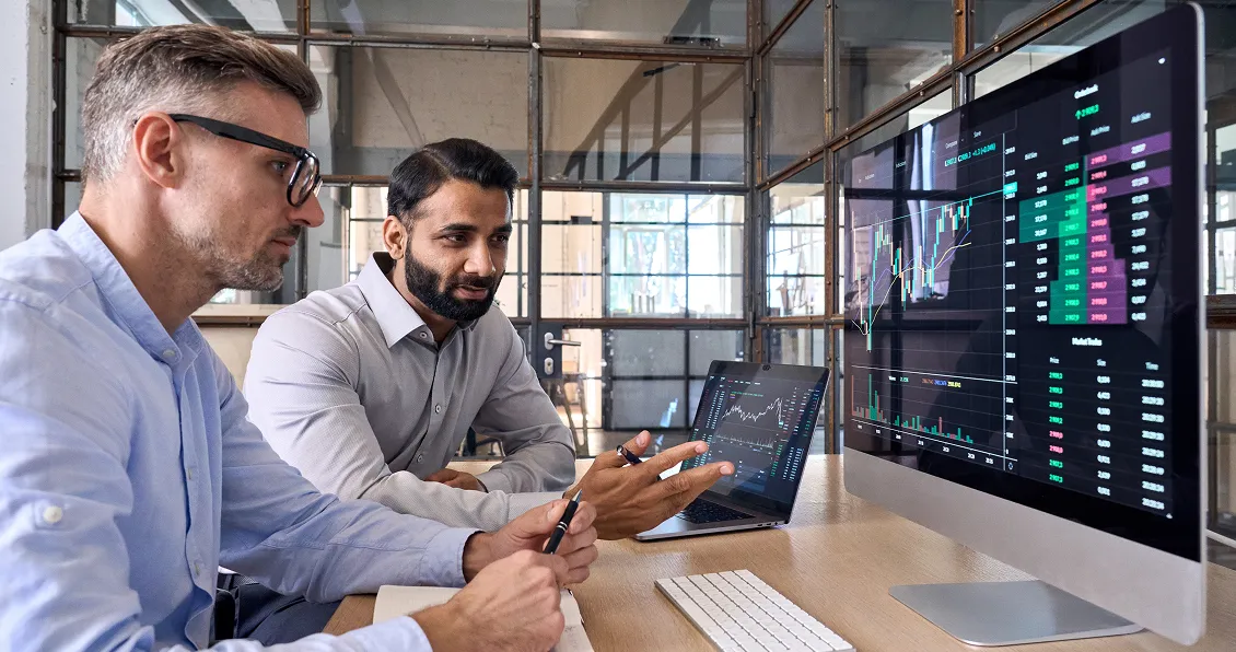 Two men in business attire sit at a desk in a modern office, analyzing financial data on a computer monitor and a laptop. Several stock charts and graphs are visible on the screens. One man gestures towards the screen while discussing with the other.