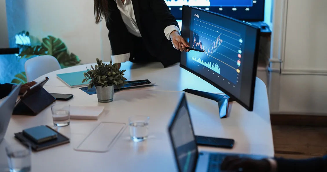 A person in business attire points at a graph on a large monitor during a meeting. The screen shows rising and falling data lines. Laptops, notebooks, water glasses, and a potted plant are on the table, with colleagues seated nearby.