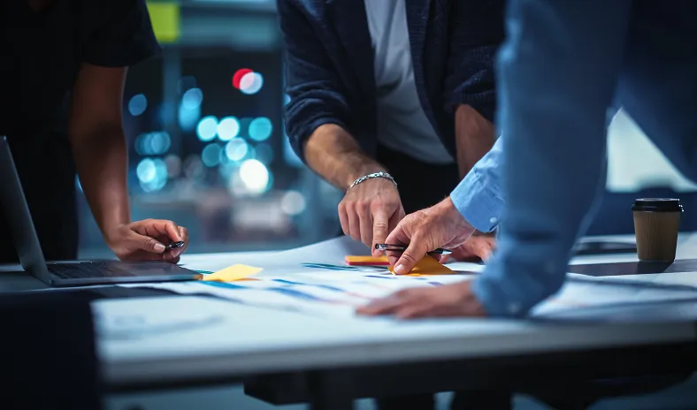 Three people, visible only from the shoulders down, lean over a table covered with papers, pens, and sticky notes, appearing to discuss or collaborate on work in a modern office setting, with blurred city lights in the background.