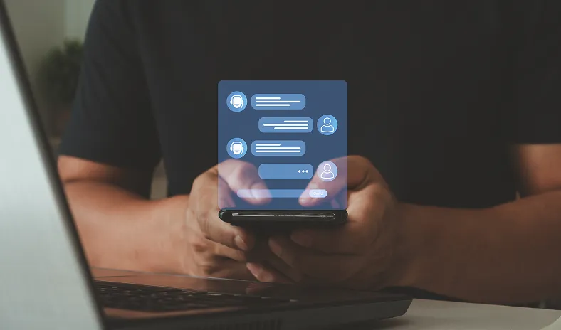 A person wearing a dark shirt holds a smartphone in both hands. In front of the phone is a virtual chat interface with speech bubbles, user icons, and chatbot symbols, suggesting messaging or a chatbot conversation. A laptop is partially visible in the foreground.