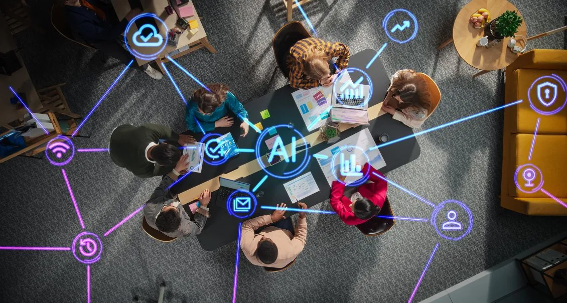 A group of people sit around a table with laptops and documents, viewed from above. Blue digital icons representing artificial intelligence and technology connect to the center of the table, emphasizing teamwork and digital collaboration.